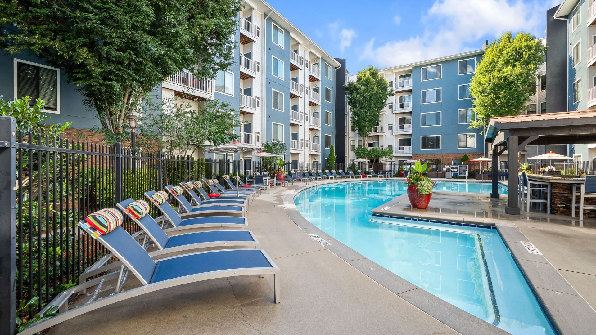 Outdoor pool area with blue lounge chairs, surrounded by apartment buildings and greenery under a blue sky.